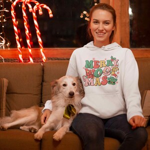 May include: A white hoodie with the text "Merry Woofmas" in colorful letters and a small elf graphic. A person is wearing the hoodie and holding a light brown dog.