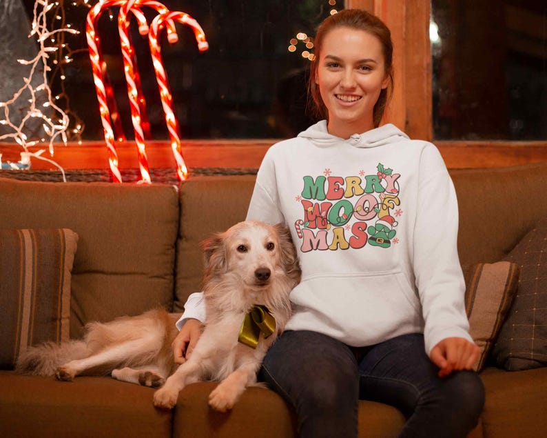 May include: A white hoodie with the text "Merry Woofmas" in colorful letters and a small elf graphic. A person is wearing the hoodie and holding a light brown dog.