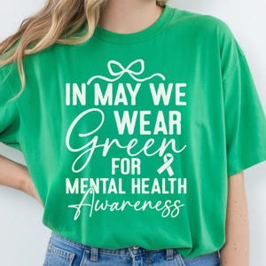 Op de afbeelding: Een felgroen t-shirt met witte tekst die het bewustzijn voor geestelijke gezondheid bevordert. De tekst luidt "IN MAY WE WEAR GREEN FOR MENTAL HEALTH Awareness" met een groen lint. Het shirt is effen van kleur en heeft een relaxte pasvorm.