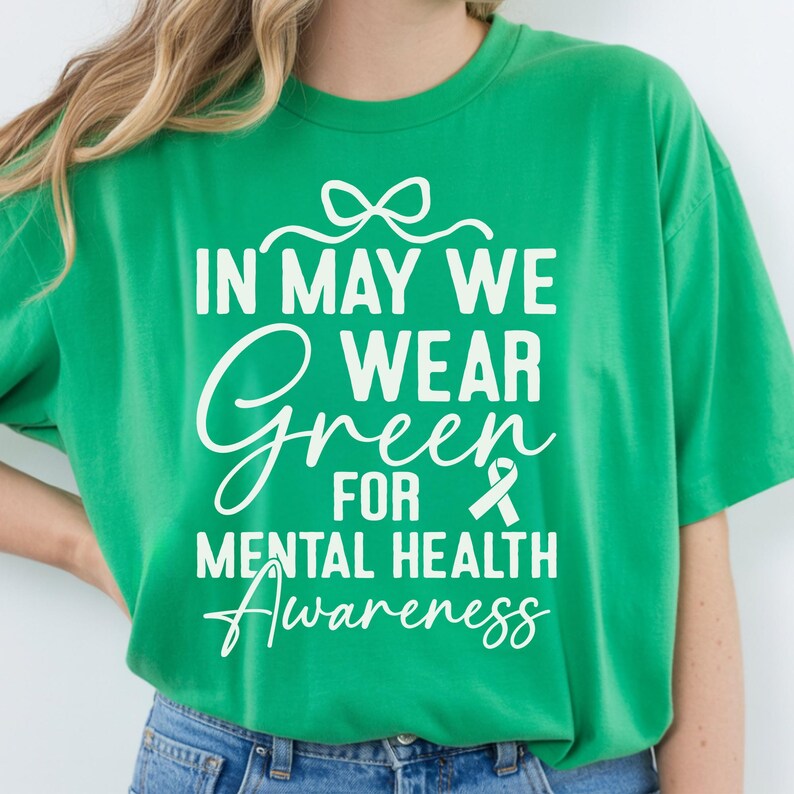 Op de afbeelding: Een felgroen t-shirt met witte tekst die het bewustzijn voor geestelijke gezondheid bevordert. De tekst luidt "IN MAY WE WEAR GREEN FOR MENTAL HEALTH Awareness" met een groen lint. Het shirt is effen van kleur en heeft een relaxte pasvorm.