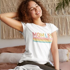 May include: White t-shirt with a retro-style graphic that says "MOMLIFE" in pink, orange, and yellow letters with colourful stripes underneath. The text "MAMA" is written in smaller letters below the stripes.