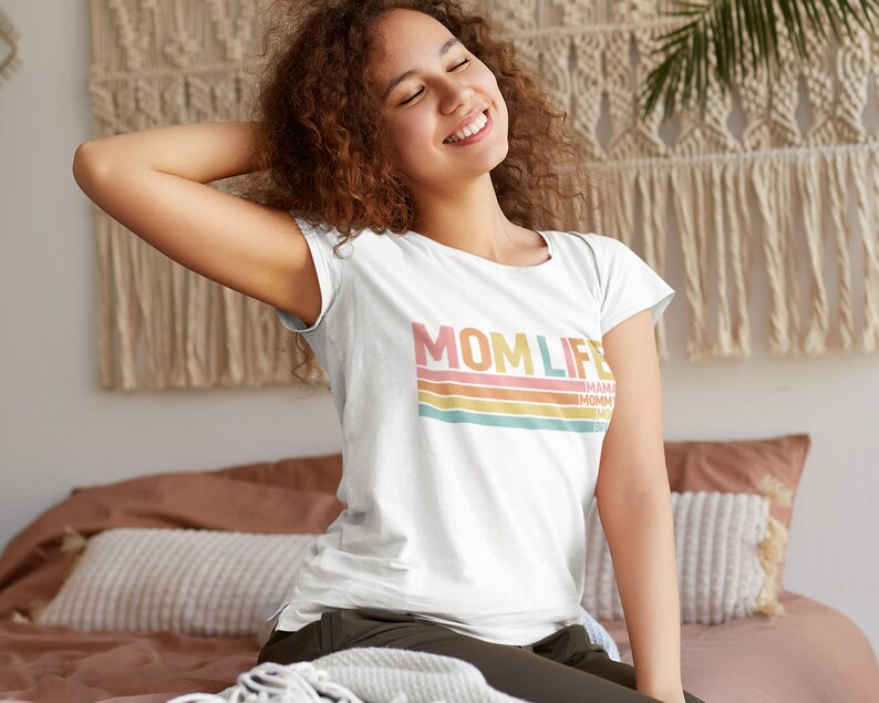 May include: White t-shirt with a retro-style graphic that says "MOMLIFE" in pink, orange, and yellow letters with colourful stripes underneath. The text "MAMA" is written in smaller letters below the stripes.