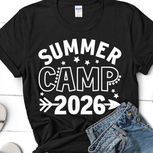 May include: Black t-shirt with the words "SUMMER CAMP 2026" in white, with stars and an arrow design. The shirt is folded with a pair of blue jeans.