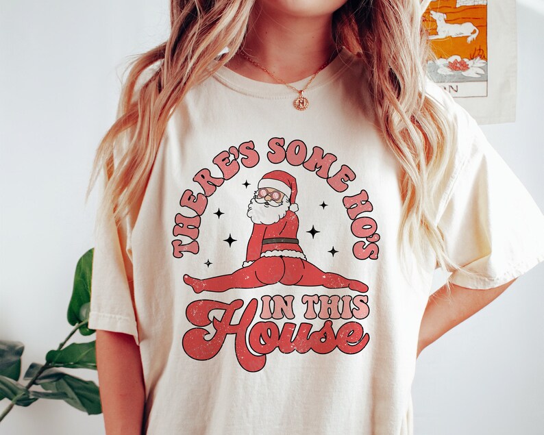 May include: A cream-colored t-shirt with a graphic of Santa Claus doing a split with the text "There's some ho's in this house".