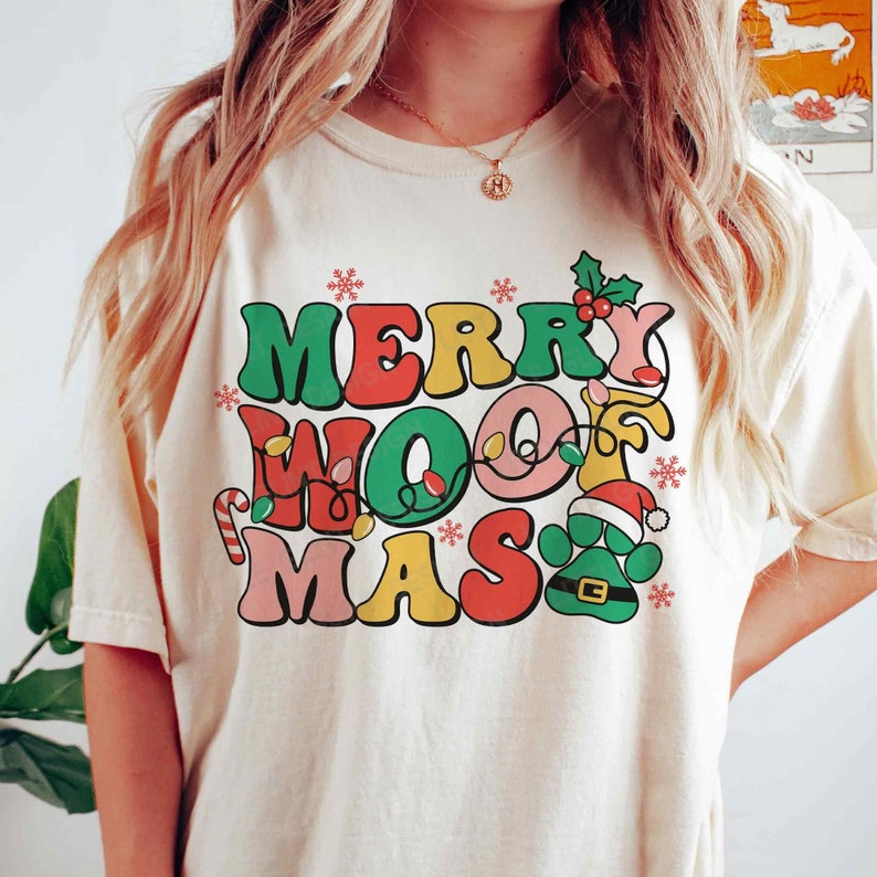 May include: A cream-colored t-shirt with the text "Merry Woof Mas" in colorful, retro-style letters. The text is decorated with Christmas lights, a candy cane, a snowflake, and a paw print wearing a Santa hat.