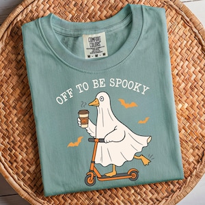 May include: A seafoam green t-shirt with the phrase "OFF TO BE SPOOKY" printed on the front. The graphic features a ghost-duck riding an orange scooter and holding a coffee cup with a pumpkin design. Orange bat silhouettes are also included.