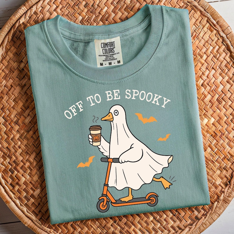 May include: A seafoam green t-shirt with the phrase "OFF TO BE SPOOKY" printed on the front. The graphic features a ghost-duck riding an orange scooter and holding a coffee cup with a pumpkin design. Orange bat silhouettes are also included.