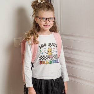 May include: A white long-sleeved shirt with a colorful graphic of a pencil with the text "100 Days Sharper". The girl is wearing a pink backpack and a black skirt.