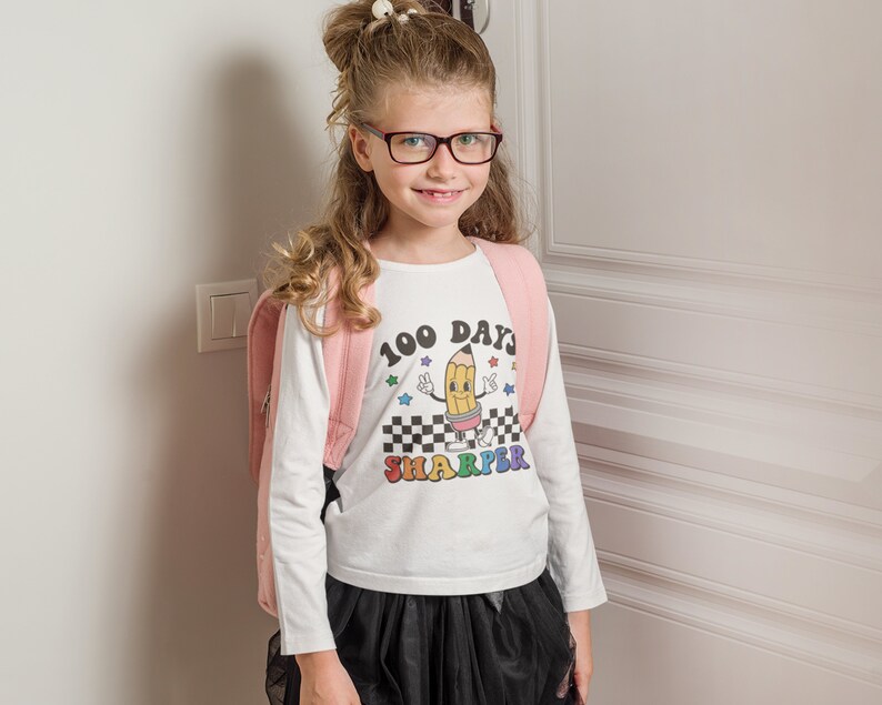 May include: A white long-sleeved shirt with a colorful graphic of a pencil with the text "100 Days Sharper". The girl is wearing a pink backpack and a black skirt.