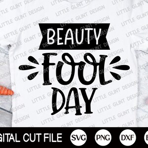 Op de afbeelding: Een wit T-shirt met zwarte tekst die "Beauty Fool Day" luidt. De tekst is in een gestileerd lettertype met een decoratieve versiering. Het shirt ligt op een houten oppervlak met een wortel en een roze ei.