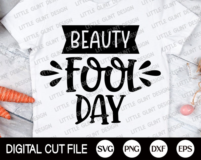 Op de afbeelding: Een wit T-shirt met zwarte tekst die "Beauty Fool Day" luidt. De tekst is in een gestileerd lettertype met een decoratieve versiering. Het shirt ligt op een houten oppervlak met een wortel en een roze ei.