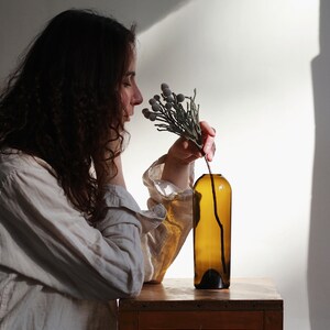 May include: A person in a white shirt is smelling a sprig of dried flowers in a brown glass bottle. The bottle is sitting on a wooden table.