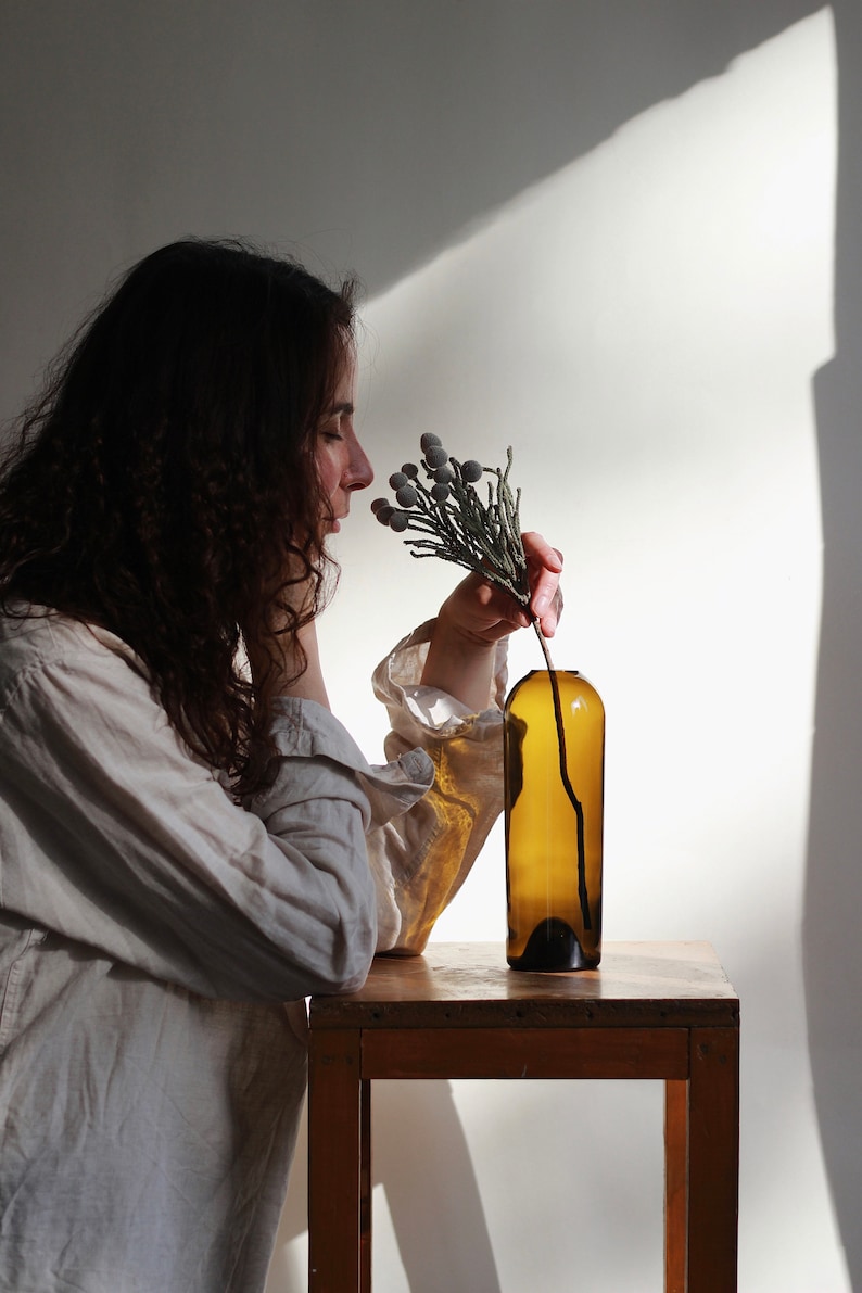 May include: A person in a white shirt is smelling a sprig of dried flowers in a brown glass bottle. The bottle is sitting on a wooden table.