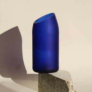 Pu&ograve; includere: Un vaso in vetro blu cobalto con un'apertura inclinata poggia su una pietra grigia ruvida. Il vaso &egrave; smerigliato e ha un design moderno e minimalista.