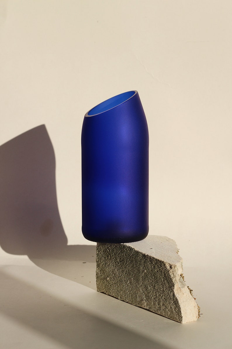 Pu&ograve; includere: Un vaso in vetro blu cobalto con un'apertura inclinata poggia su una pietra grigia ruvida. Il vaso &egrave; smerigliato e ha un design moderno e minimalista.