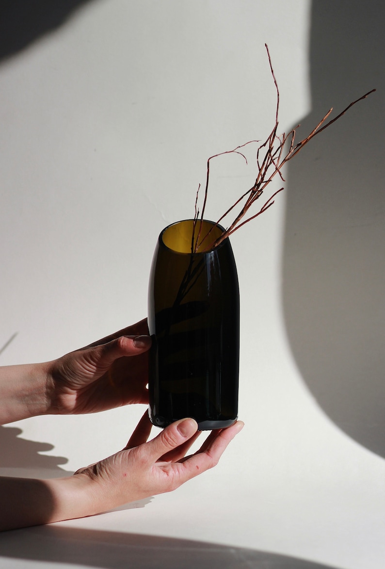 Pu&ograve; includere: Un vaso di vetro marrone scuro con un'apertura stretta, che contiene alcuni rami sottili marroni. Il vaso &egrave; tenuto da due mani.