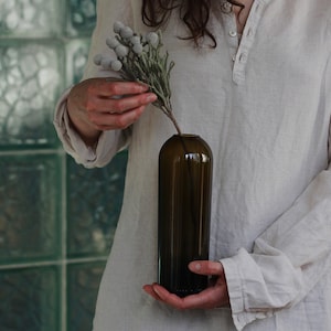 May include: A person holding a dark brown glass vase with a sprig of white and green dried flowers.
