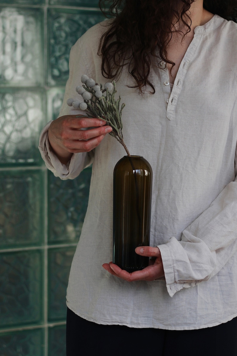 May include: A person holding a dark brown glass vase with a sprig of white and green dried flowers.