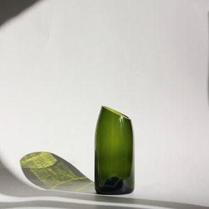 Pu&ograve; includere: Un vaso di vetro verde scuro con un'apertura angolare unica. Il vaso &egrave; appoggiato su una superficie bianca.