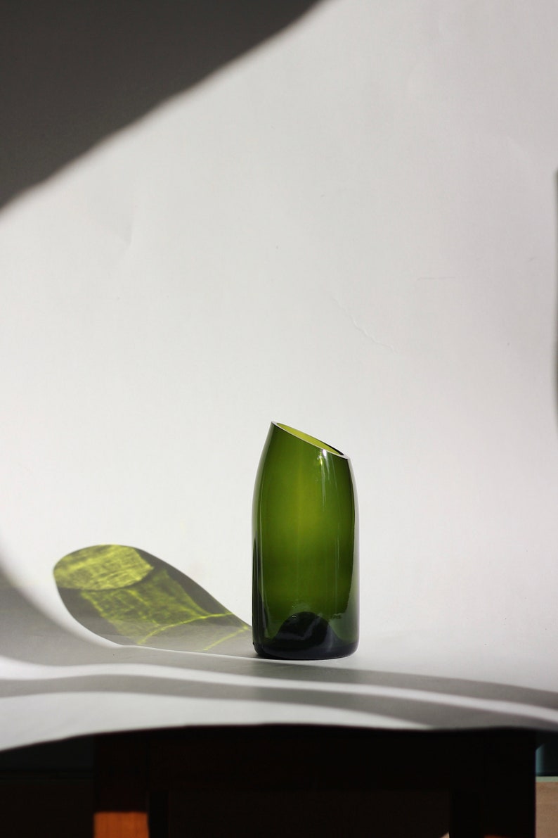 Pu&ograve; includere: Un vaso di vetro verde scuro con un'apertura angolare unica. Il vaso &egrave; appoggiato su una superficie bianca.