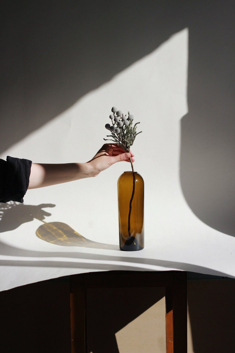 May include: A hand holds a sprig of dried, gray-green flowers with small, round, gray buds. The flowers are being placed into a brown glass bottle vase.