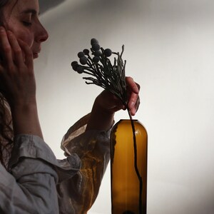 May include: A person is holding a sprig of dried flowers with gray berries and placing them into a brown glass bottle.