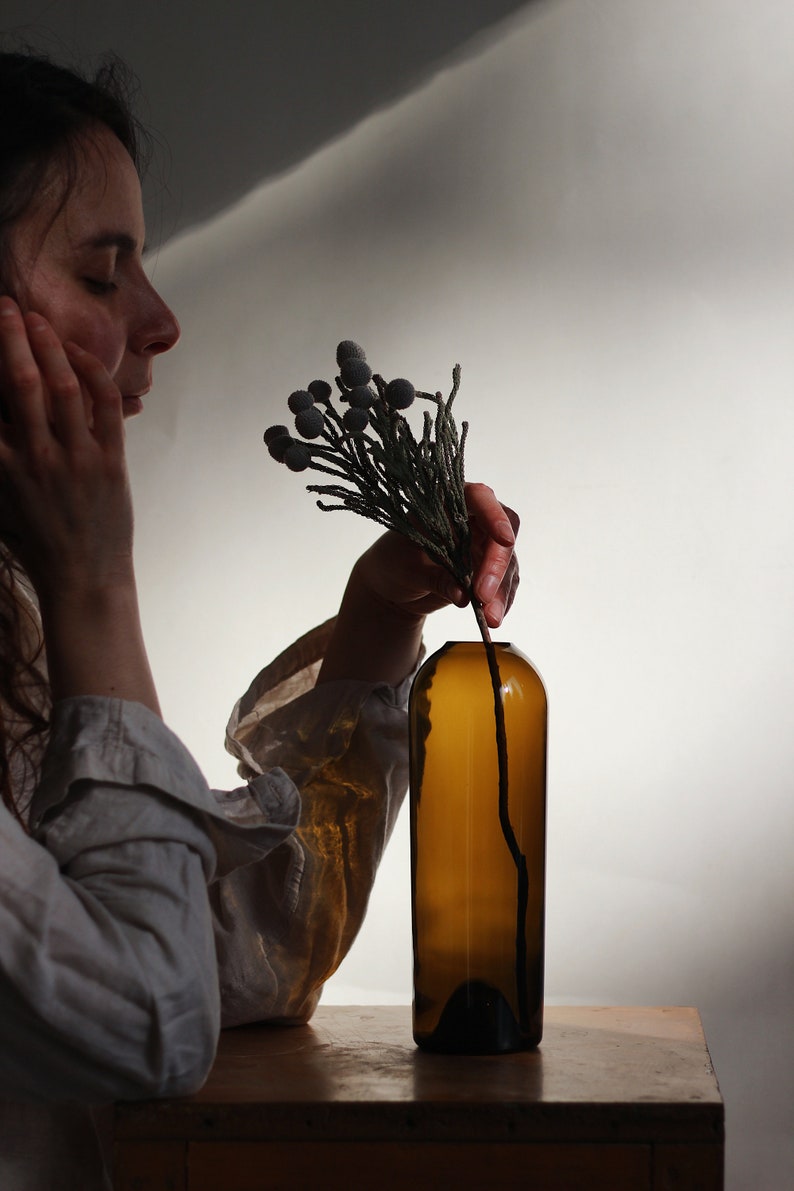 May include: A person is holding a sprig of dried flowers with gray berries and placing them into a brown glass bottle.