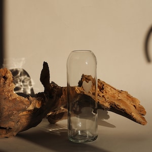 May include: A clear glass vase with a heart-shaped design sits on a white surface next to a piece of driftwood.
