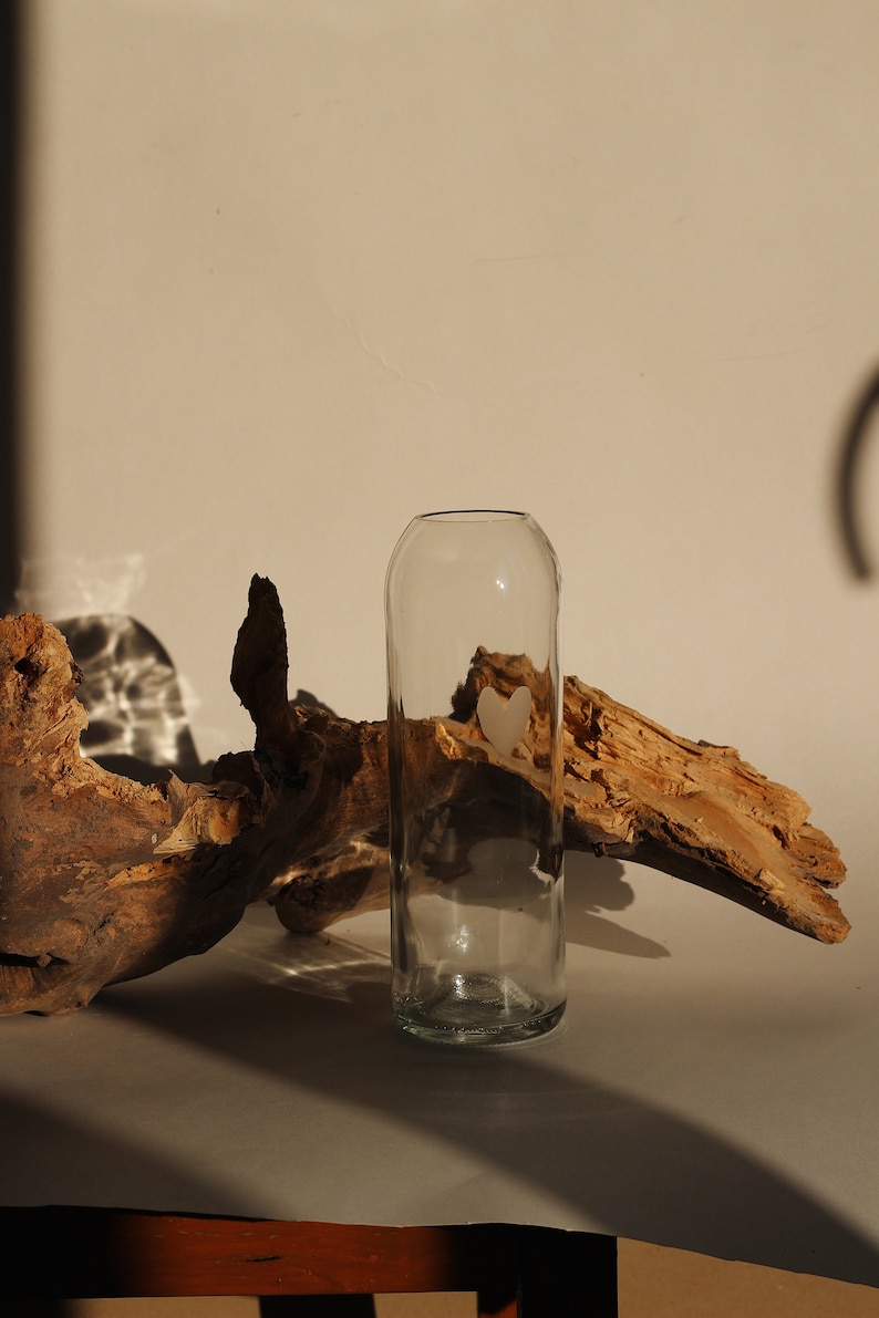 May include: A clear glass vase with a heart-shaped design sits on a white surface next to a piece of driftwood.