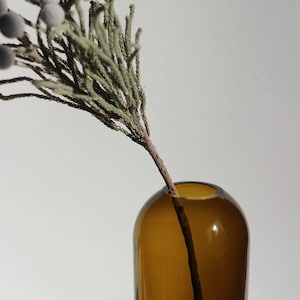 May include: A brown glass vase with a single sprig of dried greenery with small white berries.