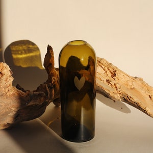 May include: A dark green glass vase with a heart etched into the side. The vase is sitting on a light brown wooden surface.