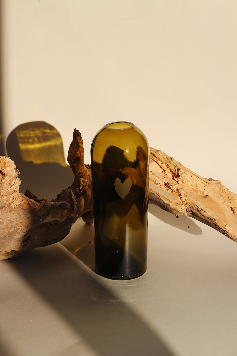 May include: A dark green glass vase with a heart etched into the side. The vase is sitting on a light brown wooden surface.