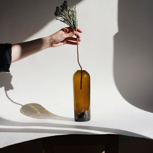 May include: A hand holds a dried flower sprig with small, round, gray buds. The sprig is placed in a brown glass bottle vase on a white surface.