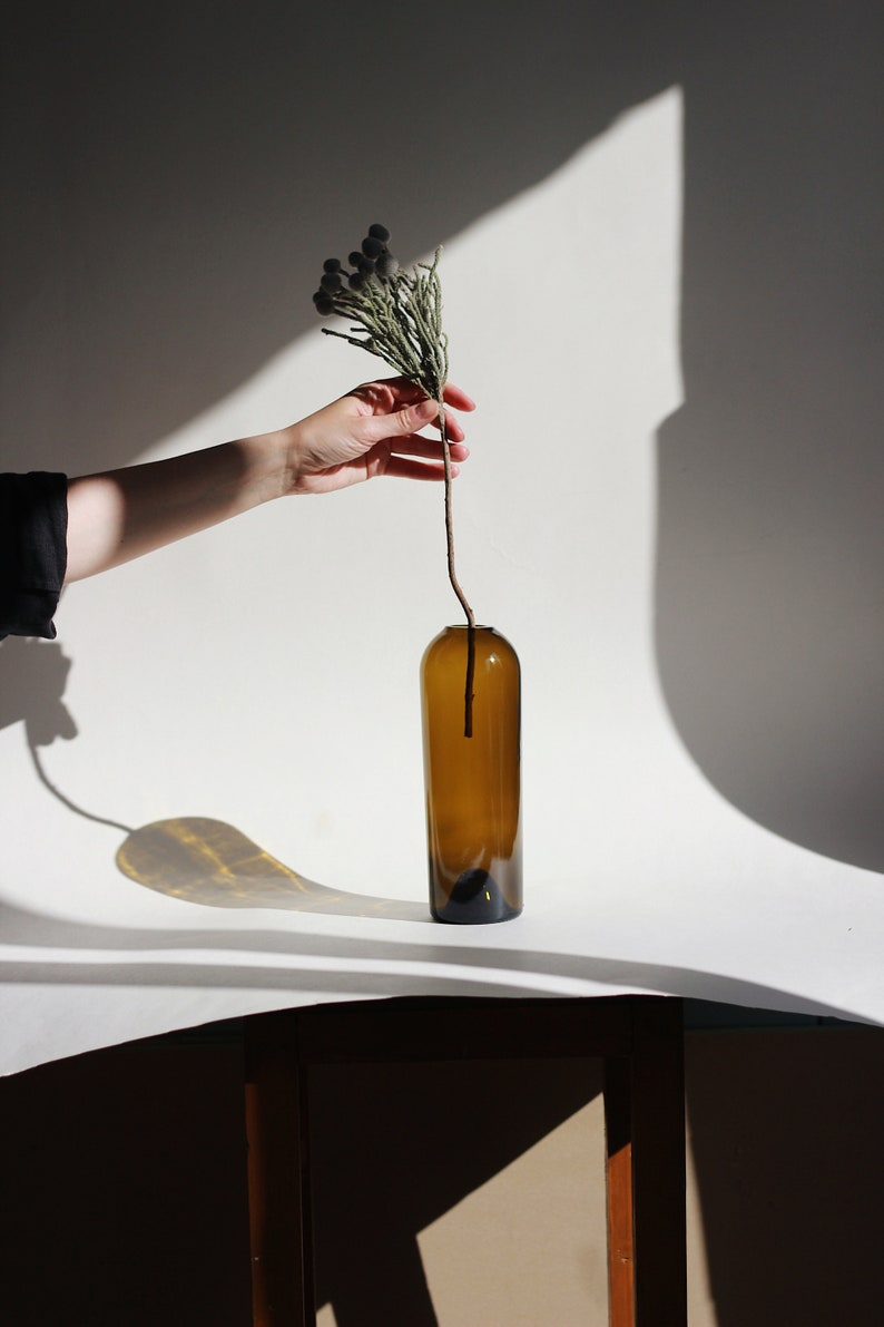 May include: A hand holds a dried flower sprig with small, round, gray buds. The sprig is placed in a brown glass bottle vase on a white surface.