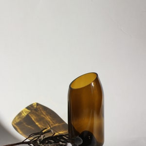 Pu&ograve; includere: Un vaso di vetro marrone con un'apertura angolare unica, posto su una superficie bianca con un ramoscello di fiori secchi.