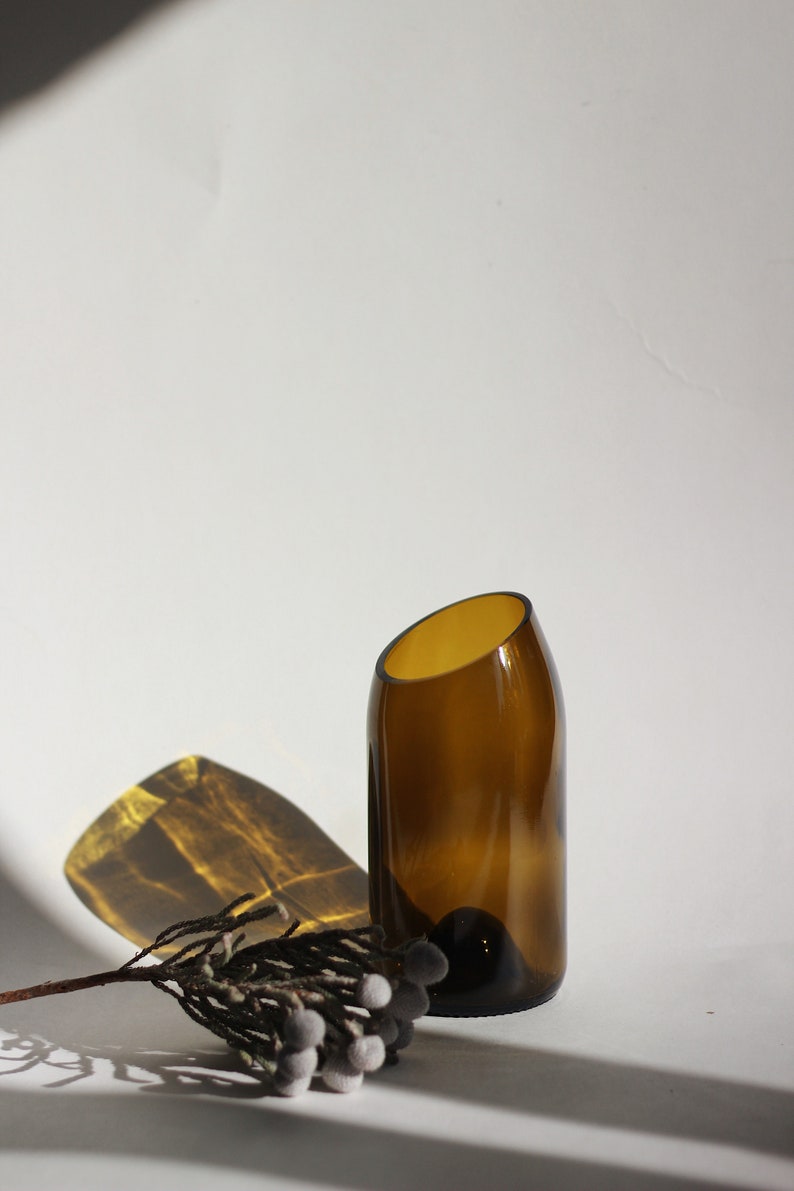 Pu&ograve; includere: Un vaso di vetro marrone con un'apertura angolare unica, posto su una superficie bianca con un ramoscello di fiori secchi.