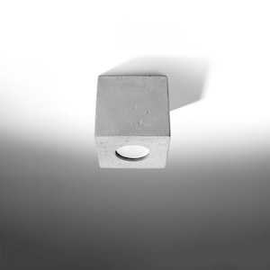 May include: A gray concrete cube-shaped light fixture with a circular opening. The light fixture is mounted on a white surface, casting a shadow. The design is minimalist and modern.