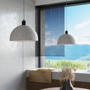 May include: Two concrete pendant lights hang over a dining table with a wooden top. The lights are suspended from black cords and have a speckled gray finish. The table is set with a bowl of quail eggs and a vase of dried flowers.