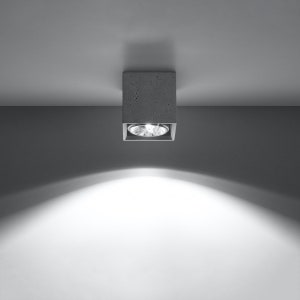 Könnte beinhalten: Eine quadratische, graue Einbau-Deckenleuchte. Das Licht ist in einem betonartigen Material eingeschlossen und strahlt einen hellen, fokussierten Lichtstrahl aus. Die Leuchte ist an einer weißen Decke montiert, mit einem Schatten an der Wand.