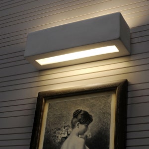 Puede incluir: Una lámpara de pared rectangular blanca con un panel empotrado iluminado. La luz está montada encima de un retrato enmarcado de una mujer. La pared tiene paneles horizontales de color claro.