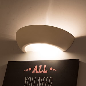 May include: A cream-colored, curved wall sconce emitting warm light. Below, a dark sign displays the text "...ALL... YOU NEED" in a pink, handwritten style font. The sconce is mounted on a white wall.