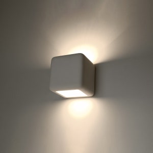 May include: A square, white wall-mounted light fixture with rounded corners. The fixture emits a bright, warm light from a recessed square opening on the bottom. The light casts a shadow on the wall.