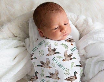 May include: A white swaddle with a repeating pattern of green leaves and flying ducks. The text "Ryan Hunter" is repeated throughout the pattern. A baby is swaddled in the blanket.