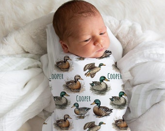 May include: A white swaddle with a pattern of brown and green ducks. The text "COOPER" is repeated throughout the swaddle. A baby is wrapped in the swaddle.