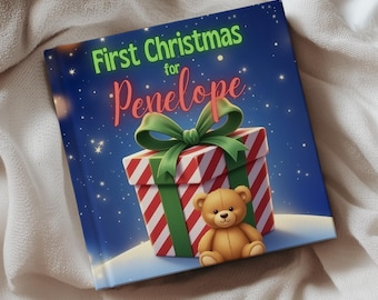 May include: A children's book titled "First Christmas for Penelope" features a gift box with a green bow and a teddy bear. The book has a blue background with stars and is resting on a white blanket.
