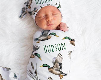 May include: A white swaddle blanket with a pattern of green and brown ducks in flight. The blanket is wrapped around a baby wearing a white hat with the name "HUDSON" written on it. The blanket also has the name "HUDSON" written on it.
