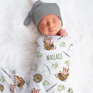 May include: A white swaddle blanket with a repeating pattern of Viking ships, swords, bows and arrows, and green leaves. The blanket has the name "Wallace" printed on it. A baby is swaddled in the blanket and is wearing a gray knitted hat.