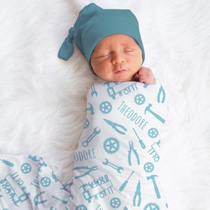 May include: A white swaddle blanket with a blue and white pattern of tools, gears, and the name "Theodore" repeated throughout. A baby is swaddled in the blanket and is wearing a blue knit hat.