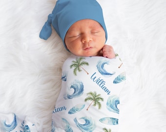 Puede incluir: Una manta de arrullo blanca con un patrón de acuarela azul y verde de olas, palmeras y tablas de surf. La manta está envuelta alrededor de un bebé que lleva un gorro azul de punto. El nombre "William" está escrito en la manta.