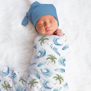 May include: A white swaddle blanket with a blue and green watercolor pattern of waves, palm trees, and surfboards. The blanket is wrapped around a baby wearing a blue knit hat. The name "William" is written on the blanket.